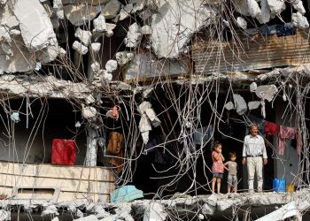 Seorang pria Palestina dan beberapa anak berdiri di depan bangunan yang rusak berat, dikelilingi besi-besi beton yang terbuka dan puing-puing, di Kota Gaza [Reuters/Mahmoud Issa]
