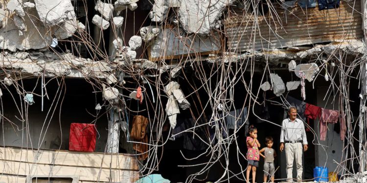 Seorang pria Palestina dan beberapa anak berdiri di depan bangunan yang rusak berat, dikelilingi besi-besi beton yang terbuka dan puing-puing, di Kota Gaza [Reuters/Mahmoud Issa]