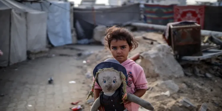 Seorang gadis Palestina yang terusir memegang boneka sambil bermain di luar tenda keluarganya di kamp pengungsian dekat pelabuhan di Kota Gaza, pada 19 Oktober 2025. [Foto oleh Majdi Fathi/NurPhoto via Getty Images]