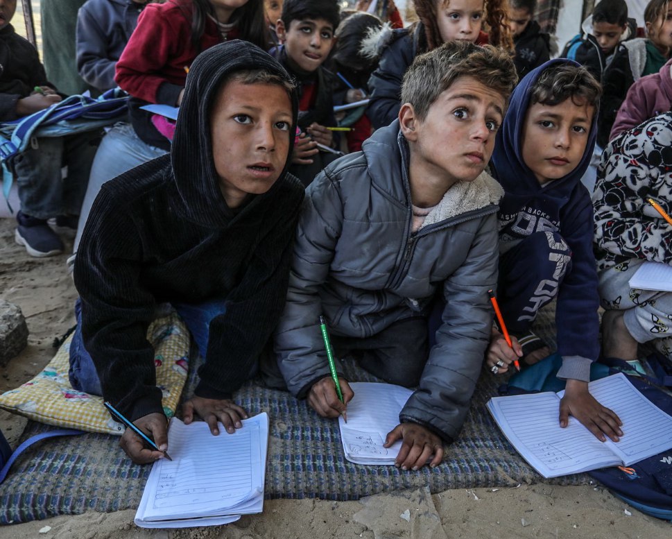 Anak-anak Palestina, yang pendidikannya terhenti akibat pengeboman Israel yang menghancurkan sekolah mereka, melanjutkan pelajaran mereka di tenda-tenda darurat bersama guru-guru sukarelawan di Khan Younis (18/10). Sebanyak 97% sekolah hancur atau rusak, mengakibatkan 600.000 anak usia sekolah memulai tahun ketiga mereka tanpa tempat belajar yang layak. (Abed Rahim Khatib/Anadolu/The Guardian)