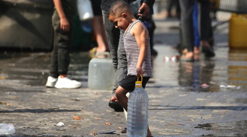 Anak Gaza membawa botol air di tanah yang basah dan kotor oleh air hujan (MEE)