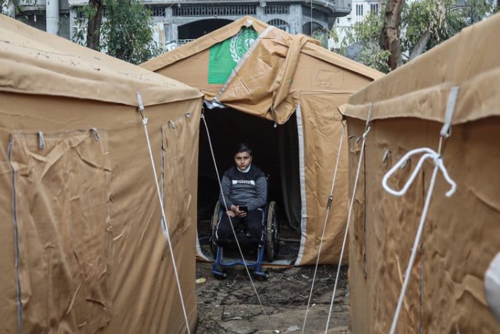 Seorang anak difabel duduk di kursi roda di antara tenda pengungsian Gaza (Mondoweiss)