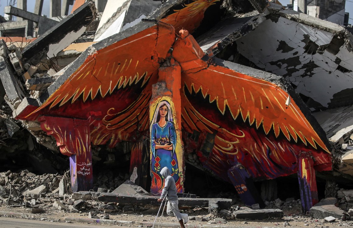 Seorang pria yang menggunakan tongkat berjalan melewati mural di sisi bangunan yang hancur di Kamp Pengungsi Maghazi di Deir al-Balah (7/12). Karya ini ditafsirkan oleh sebagian orang sebagai simbol keteguhan, sementara simbol burung phoenix yang bangkit dari puing-puing melambangkan ketahanan dan harapan di Gaza. (Eyad Baba/AFP/ The Guardian)