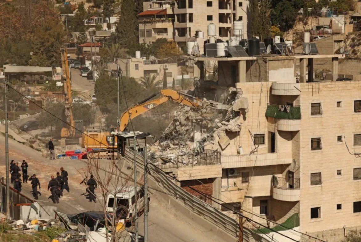 Pasukan Israel berkumpul saat ekskavator menghancurkan sebuah bangunan Palestina di dekat lingkungan Silwan di Yerusalem bagian timur (22/12). Mereka juga menghancurkan toko-toko di sekitar Kamp Pengungsi Qalandiya, serta memperluas penggerebekan ke Kota Kafr Aqab untuk melakukan penggeledahan rumah dan secara paksa mengusir penduduk dari rumah mereka. (AFP/ Al-Jazeera)