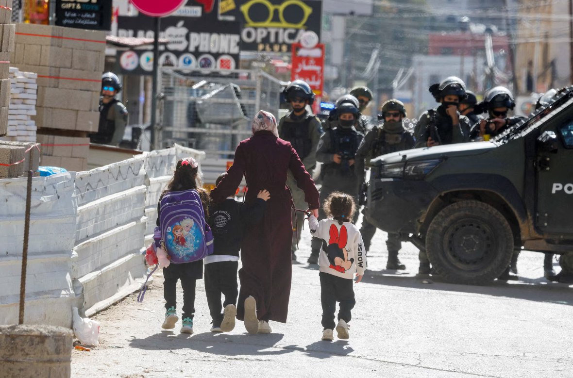Seorang ibu Palestina bersama anak-anaknya berjalan di dekat anggota polisi perbatasan Israel selama penggerebekan Israel di Qalandia, Tepi Barat yang diduduki (26/1). Pasukan Israel melancarkan agresi militer skala besar di sekitar kamp pengungsi Qalandia, menghancurkan bangunan milik Palestina, menilang sejumlah besar kendaraan warga, dan merusak kendaraan lainnya. (Mohamad Torokman/ Reuters/ WAFA/ Anadolu)